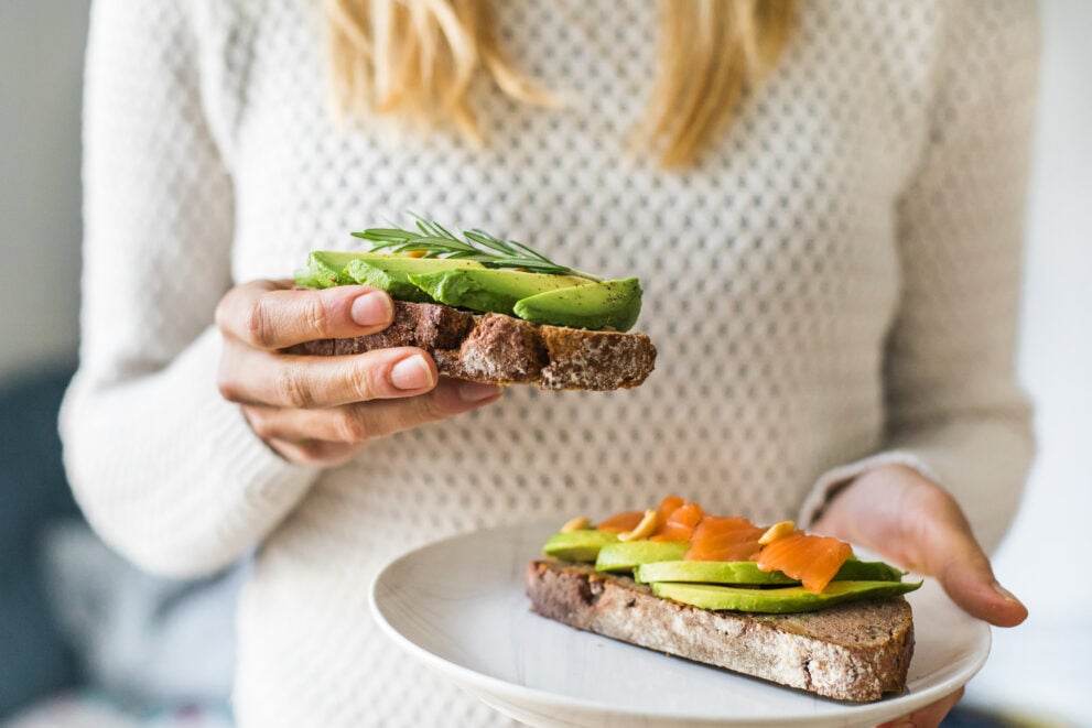 A woman is about to eat piece of toast. The toast has avocado and salmon on top.