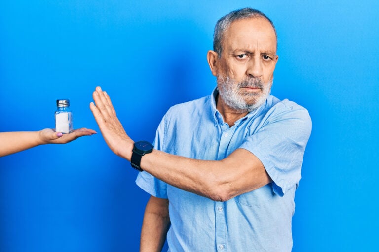A senior man with a beard is being offering a salt shaker. He has an open hand doing a stop gesture with serious and confident expression