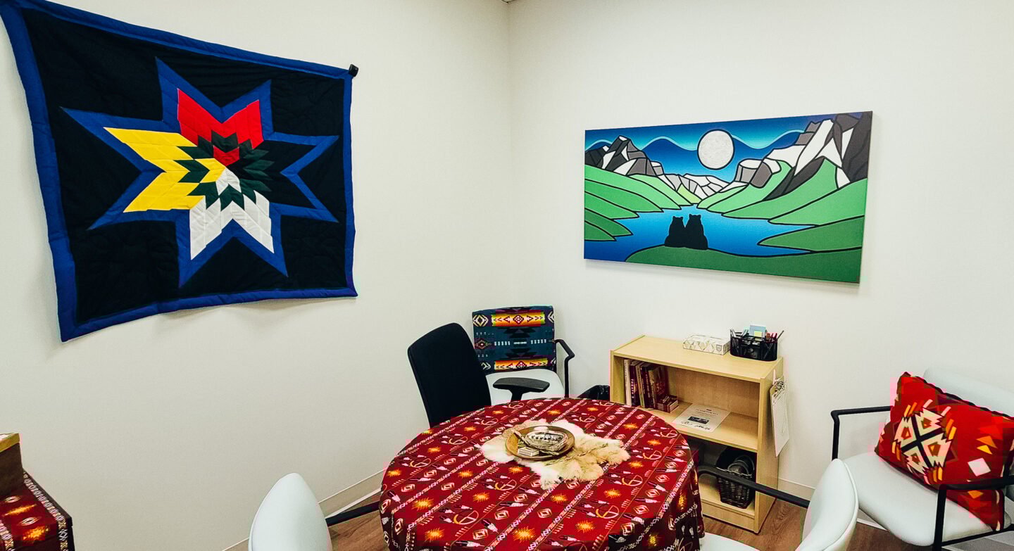 A wide shot of the smudge room at the PCN's Uxborough location. You can see Indigenous art on the walls, and a round table is in the middle of the room with smudging materials on top.