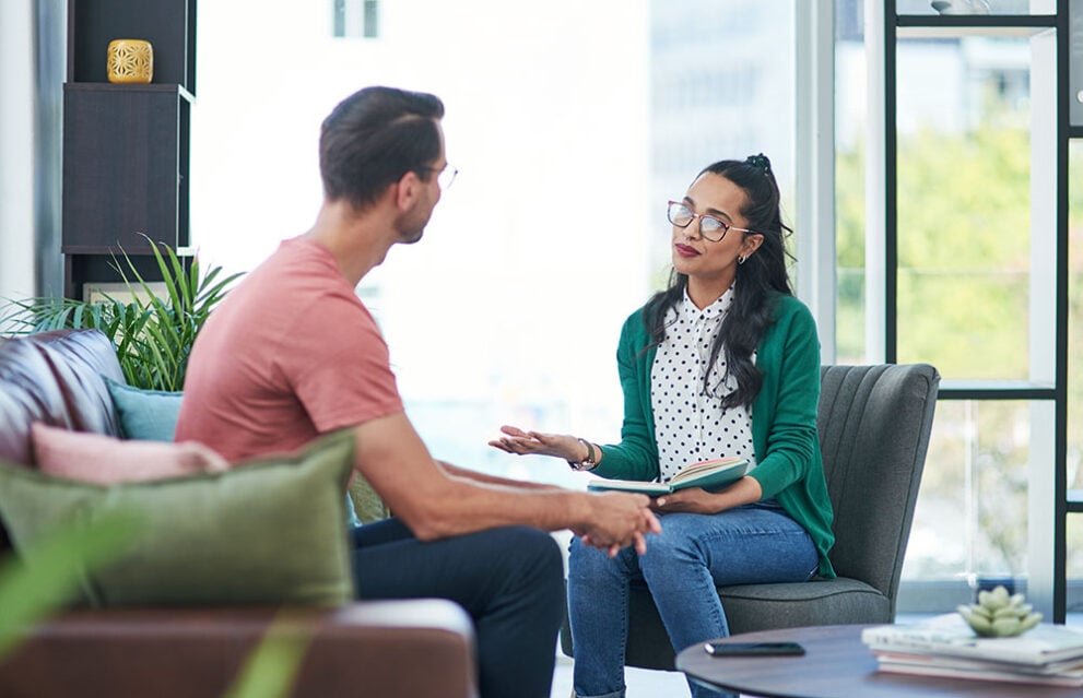 A man and a woman sitting across from one another, having a conversation. The woman is holding a notebook and is providing counselling the man.