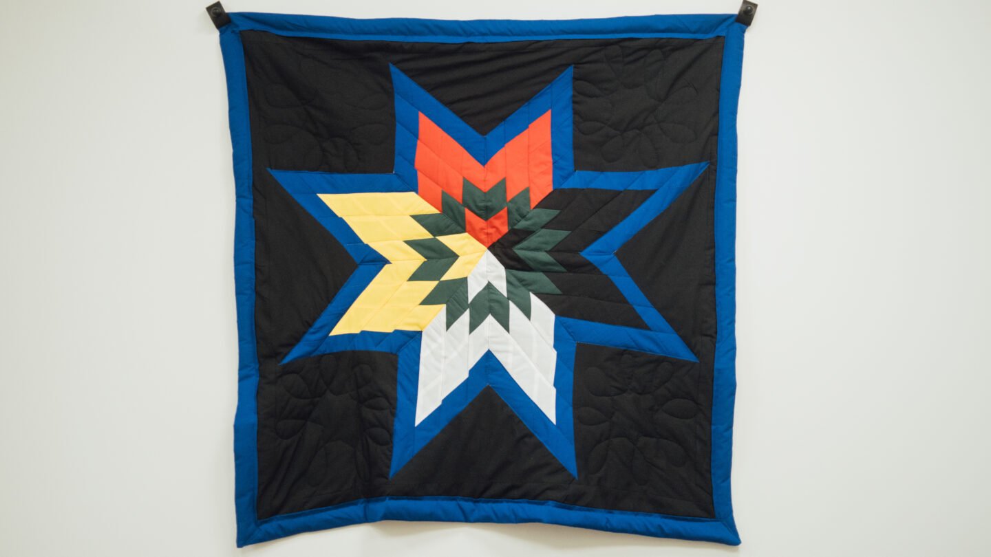 A quilted blanket hangs on the wall. The quilt is black with a star in the middle. The star is coloured to align with the traditional medicine wheel: red, yellow, black, and white.