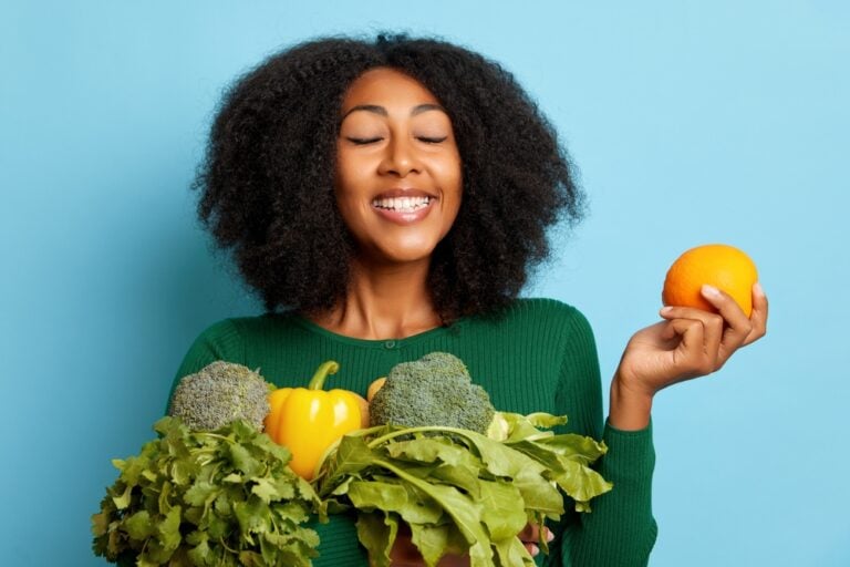 A young woman holds a bag of vegetables against a blue background. She is smiling.