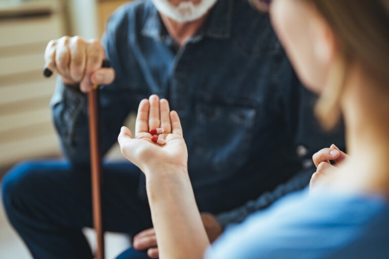 A senior patient sits with a health care provider. The health care provider shows the patient a few pills, which are resting in their palm.