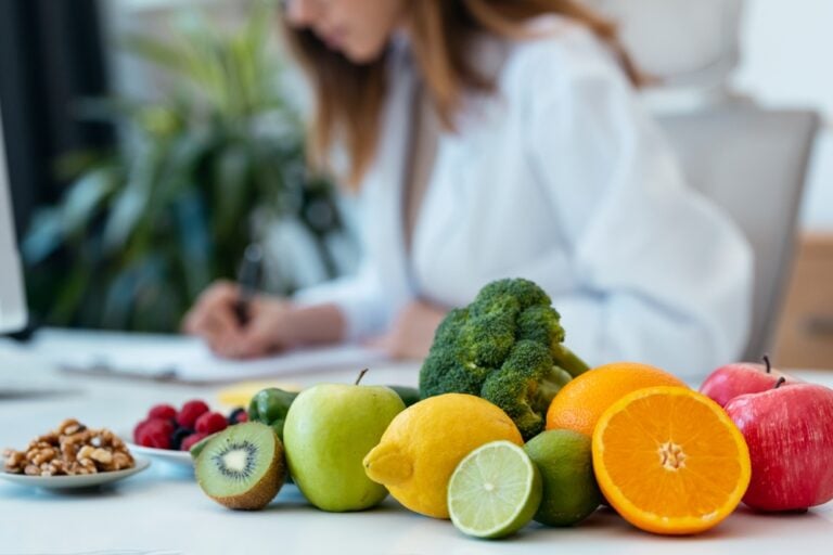 In the front, we see a mixed array of colourful fruits and vegetables. In the distance, we see a female dietitian writing notes.