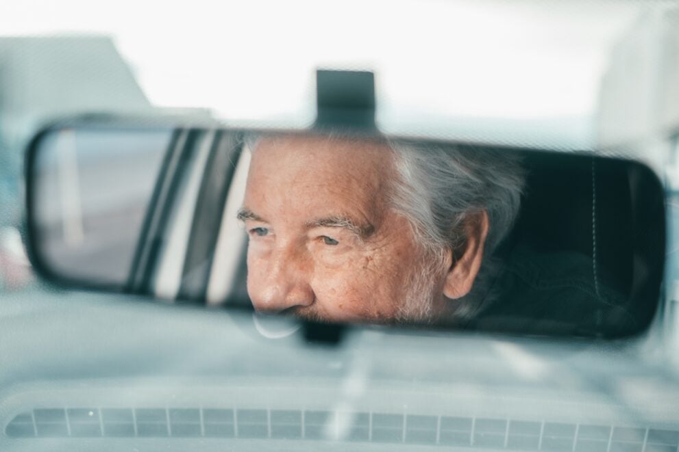We see a senior man driving a car. We see his eyes looking forward through the rearview mirror.