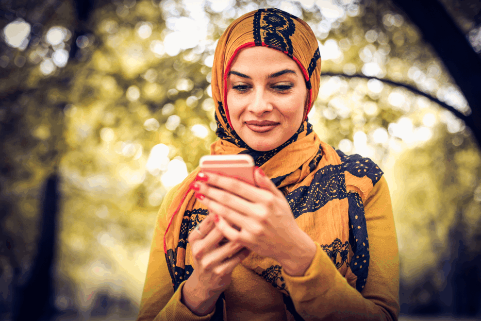 A photo of a woman in a headscarf, holding her mobile device. The background is a blurred forest scene