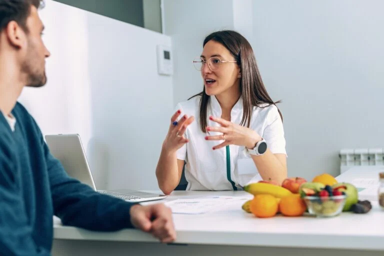 A female dietitian speaks to a male patient with animated hands.