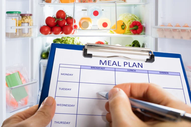 A person's hand is seen filling in a 'Meal Plan' document on a clipboard for the week. They are standing in front of an open fridge.
