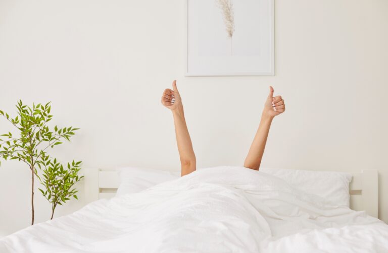 Female person waking up in the morning lying in bed under cozy white blanket in bedroom, stretching arms and showing thumb up sign. Relaxed sleepy woman happy to start a new day in the morning.