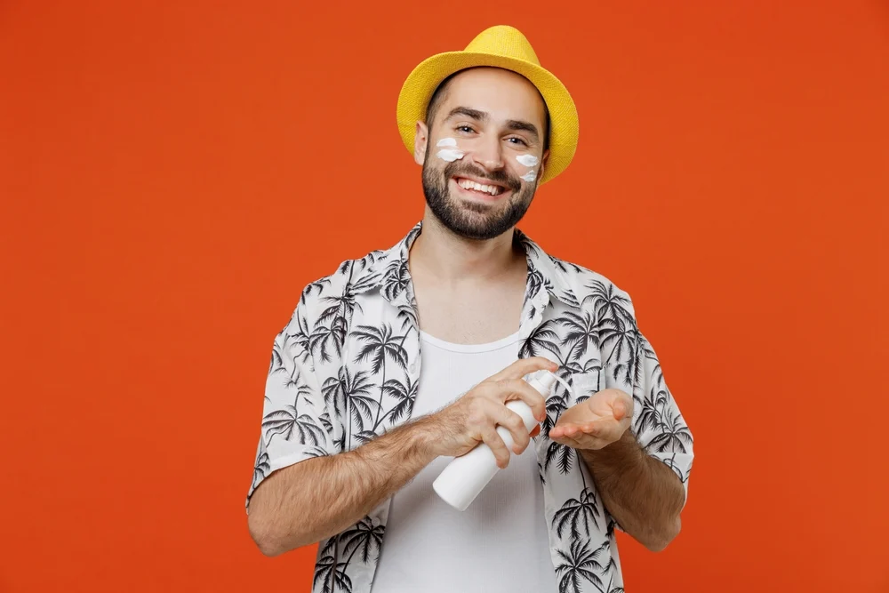 A man wearing a sunhat and tropical shirt smiles while he applies sunscreen.